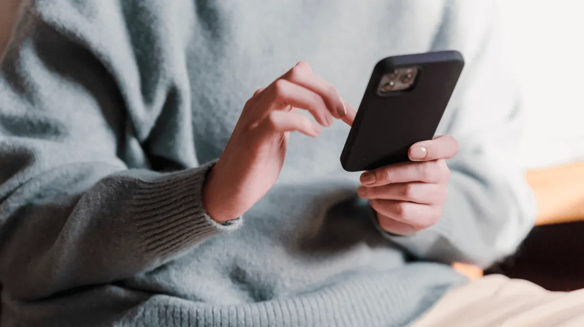 Osoba w jasnym swetrze korzysta z telefonu, siedząc w domu